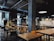 A modern industrial-style cafe with exposed ceilings and concrete pillars. There are several wooden tables and yellow chairs neatly arranged throughout the space. Hanging lights provide illumination, and plants are positioned strategically around the room. In the background, there is a tiled counter area with menus written on blackboards.