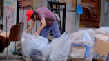 An elderly person wearing a red cap and striped clothing is sorting through several large transparent bags filled with recyclables and cardboard boxes on a city sidewalk. Surrounding the person are signs in another language, some depicting services offered inside nearby establishments.