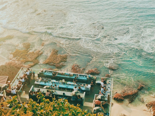 A beautiful seaside bar perched on the edge of a coastline, overlooking turquoise waters with visible coral reef formations. Groups of people are gathered around a stylish outdoor bar, enjoying drinks and socializing. Lush greenery frames the lower part of the scene, enhancing the vibrant and relaxed atmosphere.
