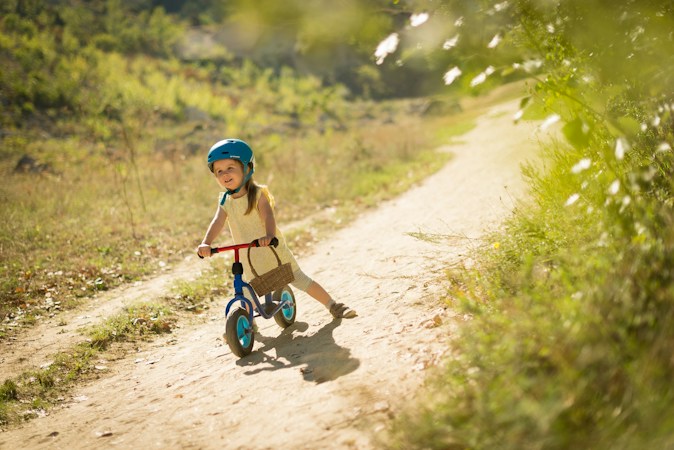 Smiling toddler in a blue helmet gliding on a small balance bike down a sunny dirt path