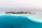 A small tropical island surrounded by turquoise blue waters. The island is densely covered with green foliage and has a few scattered buildings. There are several boats anchored in the shallow waters near the shore. On the left and right ends of the island, tall red and white communication towers are visible.