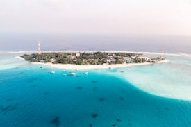 A small tropical island surrounded by turquoise blue waters. The island is densely covered with green foliage and has a few scattered buildings. There are several boats anchored in the shallow waters near the shore. On the left and right ends of the island, tall red and white communication towers are visible.