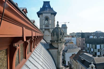 A rooftop view of an urban area featuring a historic building with ornate stone towers and decorative elements. The foreground reveals a sloped roof with red trim and arched windows. In the background, a mix of modern and historic architecture is visible, including a tower crane indicating ongoing construction.