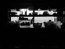 A dimly lit kitchen features shelves filled with various kitchenware silhouetted against a bright window. The shelves hold an assortment of bowls, bottles, and containers, creating a striking contrast between the dark objects and the light filtering through the window. Cutting utensils and other kitchen items hang from hooks, adding to the utilitarian aesthetic.