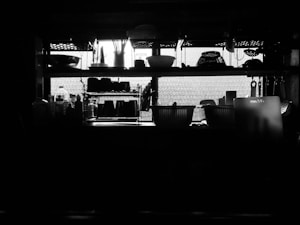 A dimly lit kitchen features shelves filled with various kitchenware silhouetted against a bright window. The shelves hold an assortment of bowls, bottles, and containers, creating a striking contrast between the dark objects and the light filtering through the window. Cutting utensils and other kitchen items hang from hooks, adding to the utilitarian aesthetic.