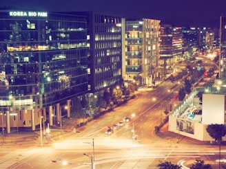 A vibrant nighttime cityscape featuring a busy street lined with modern office buildings, one labeled 'Korea Bio Park'. The scene is illuminated with streetlights, and cars are moving along the road. The buildings have reflective glass windows, showing various reflections of lights.