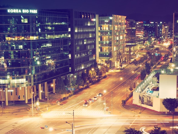 A vibrant nighttime cityscape featuring a busy street lined with modern office buildings, one labeled 'Korea Bio Park'. The scene is illuminated with streetlights, and cars are moving along the road. The buildings have reflective glass windows, showing various reflections of lights.