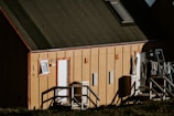 A wooden building with a sloped roof, featuring two entrances marked with numbers 368 and 369. A small window and door are present on the front facade of the building. Clothes are hanging to dry on a line outside, and there are stairs leading up to the doors.