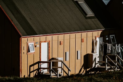 A wooden building with a sloped roof, featuring two entrances marked with numbers 368 and 369. A small window and door are present on the front facade of the building. Clothes are hanging to dry on a line outside, and there are stairs leading up to the doors.