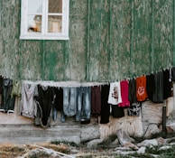 Clothes are hanging on a line against a weathered green wooden wall. Garments include shirts, pants, and dresses of various colors and styles. The building has a small window above the line.