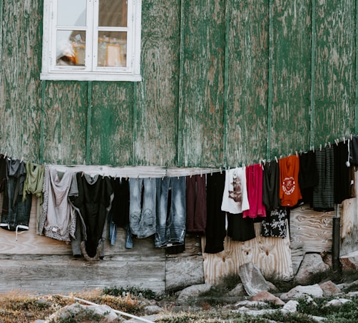 Clothes are hanging on a line against a weathered green wooden wall. Garments include shirts, pants, and dresses of various colors and styles. The building has a small window above the line.