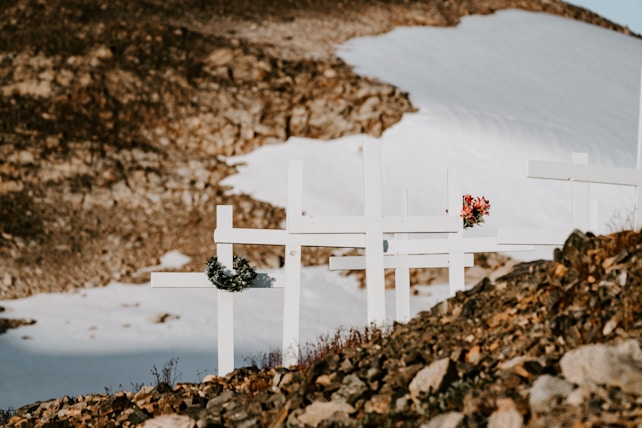 A series of white crosses with floral wreaths and bouquets are placed on a rocky terrain. In the background, there's a snow-covered mountain slope, contrasting with the brown rocky foreground.