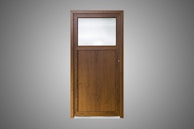 A wooden door with a simple design, featuring a large, frosted glass pane on the upper section and a solid wood panel below. The wood is of a dark brown color with a visible grain pattern. The background is a plain gray, highlighting the door.