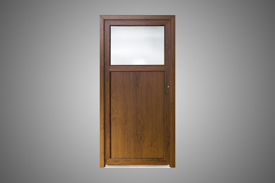 A wooden door with a simple design, featuring a large, frosted glass pane on the upper section and a solid wood panel below. The wood is of a dark brown color with a visible grain pattern. The background is a plain gray, highlighting the door.