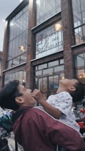 A man is joyfully holding a child in front of a large building with a sign that reads 'Tempio del Gelato'. The architecture includes large windows and brickwork illuminated by exterior lights.