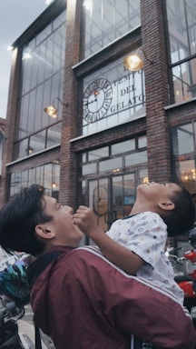 A man is joyfully holding a child in front of a large building with a sign that reads 'Tempio del Gelato'. The architecture includes large windows and brickwork illuminated by exterior lights.