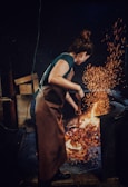 A person with brown hair wearing a brown apron is tending to a fire with a metal tool in what appears to be a workshop or forge. Bright orange sparks fly upward from the flames, illuminating the dark environment.