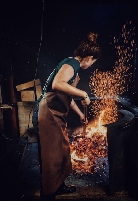 A person with brown hair wearing a brown apron is tending to a fire with a metal tool in what appears to be a workshop or forge. Bright orange sparks fly upward from the flames, illuminating the dark environment.