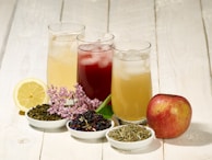 Three tall glasses filled with different types of iced tea are arranged on a rustic wooden surface. Accompanying the drinks, there is a half lemon, an apple, and small bowls filled with assorted tea leaves and herbs. A small cluster of purple flowers is also present, adding a natural decorative touch.