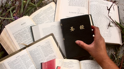 Several open books display text in different languages, including Hebrew and Latin script, spread on a surface with greenery around them. A hand holds a black book with gold lettering.