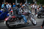 A group of motorcyclists wearing helmets are gathered on a street, surrounded by spectators. The motorcycles have shiny chrome parts and custom designs. People are casually dressed, and the scene appears to be in an urban area with buildings in the background.