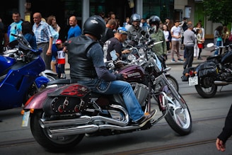 A group of motorcyclists wearing helmets are gathered on a street, surrounded by spectators. The motorcycles have shiny chrome parts and custom designs. People are casually dressed, and the scene appears to be in an urban area with buildings in the background.