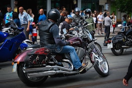 A group of motorcyclists wearing helmets are gathered on a street, surrounded by spectators. The motorcycles have shiny chrome parts and custom designs. People are casually dressed, and the scene appears to be in an urban area with buildings in the background.