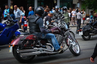A group of motorcyclists wearing helmets are gathered on a street, surrounded by spectators. The motorcycles have shiny chrome parts and custom designs. People are casually dressed, and the scene appears to be in an urban area with buildings in the background.