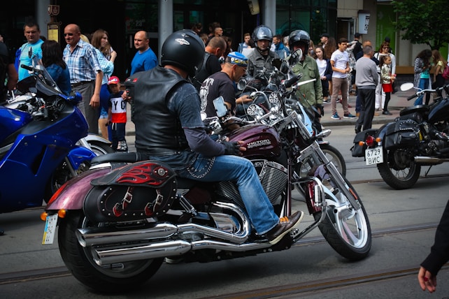 A group of motorcyclists wearing helmets are gathered on a street, surrounded by spectators. The motorcycles have shiny chrome parts and custom designs. People are casually dressed, and the scene appears to be in an urban area with buildings in the background.