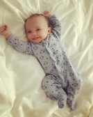 A smiling baby lies on a bed wearing a light blue onesie with cute animal patterns. The arms are raised, and the baby looks content and comfortable on the soft, white bedding.