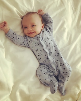 A smiling baby lies on a bed wearing a light blue onesie with cute animal patterns. The arms are raised, and the baby looks content and comfortable on the soft, white bedding.
