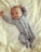 A smiling baby lies on a bed wearing a light blue onesie with cute animal patterns. The arms are raised, and the baby looks content and comfortable on the soft, white bedding.