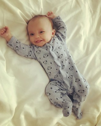 A smiling baby lies on a bed wearing a light blue onesie with cute animal patterns. The arms are raised, and the baby looks content and comfortable on the soft, white bedding.