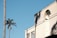 A minimalistic architectural scene featuring a pale building with geometric shapes, such as an arch and rectangular windows. Shadows cast by nearby trees are visible on the building's surface. A tall palm tree stands in the foreground against a clear blue sky, adding contrast and a hint of nature to the urban setting.