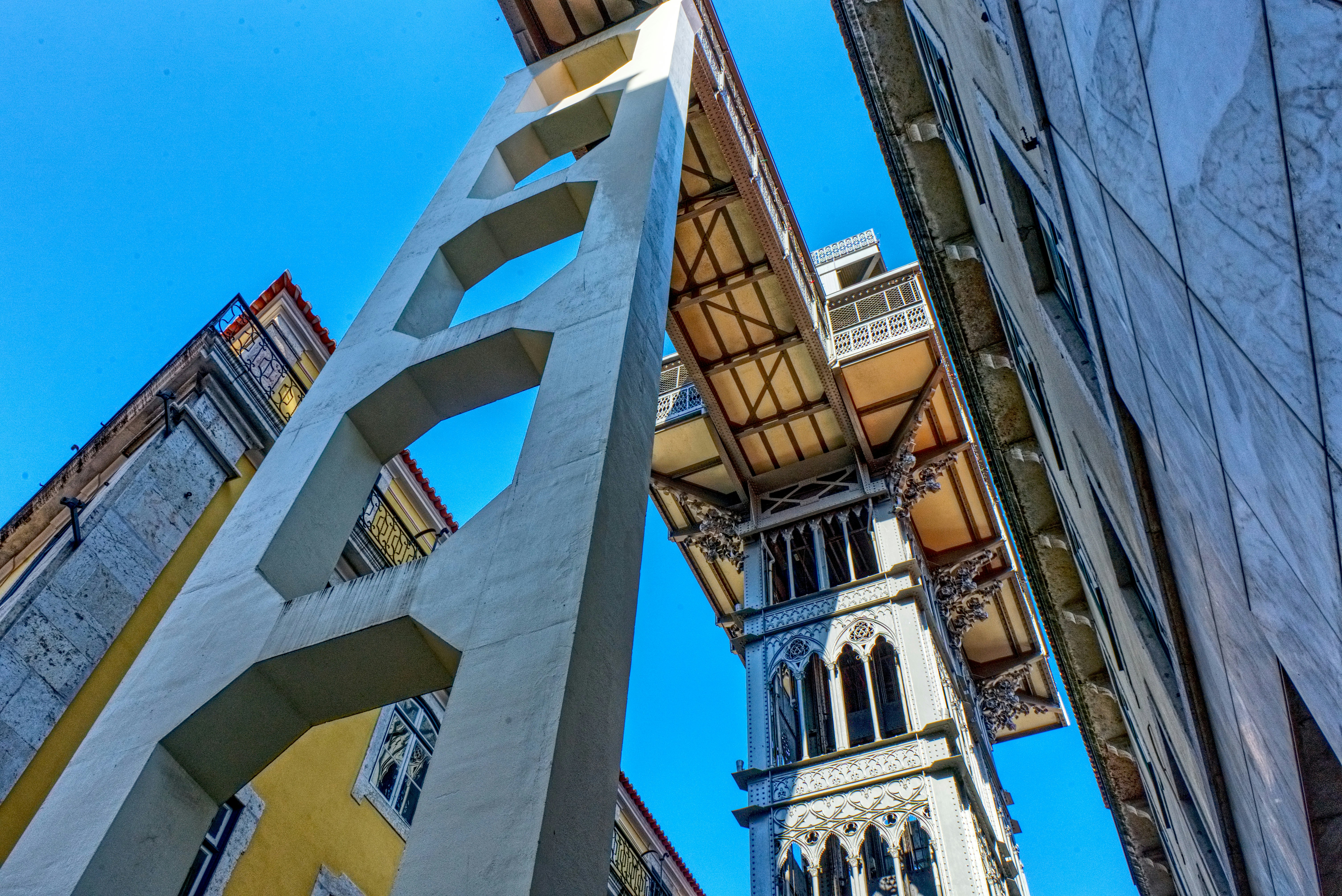 A tall, ornate metal structure resembling an elevator or lift is positioned between two buildings, with architectural details and intricate designs. The perspective is from ground level looking up, with a clear blue sky in the background and sunlight casting shadows.