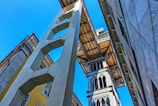 A tall, ornate metal structure resembling an elevator or lift is positioned between two buildings, with architectural details and intricate designs. The perspective is from ground level looking up, with a clear blue sky in the background and sunlight casting shadows.