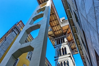 A tall, ornate metal structure resembling an elevator or lift is positioned between two buildings, with architectural details and intricate designs. The perspective is from ground level looking up, with a clear blue sky in the background and sunlight casting shadows.