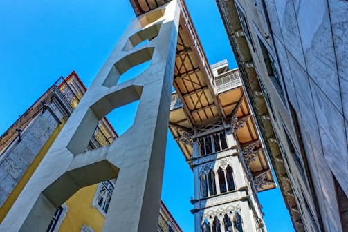 A tall, ornate metal structure resembling an elevator or lift is positioned between two buildings, with architectural details and intricate designs. The perspective is from ground level looking up, with a clear blue sky in the background and sunlight casting shadows.