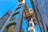 A tall, ornate metal structure resembling an elevator or lift is positioned between two buildings, with architectural details and intricate designs. The perspective is from ground level looking up, with a clear blue sky in the background and sunlight casting shadows.