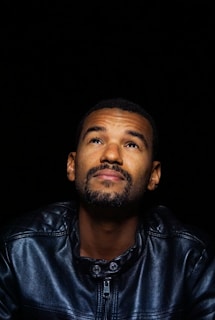 A man with a beard is looking upwards against a black background, wearing a leather jacket.