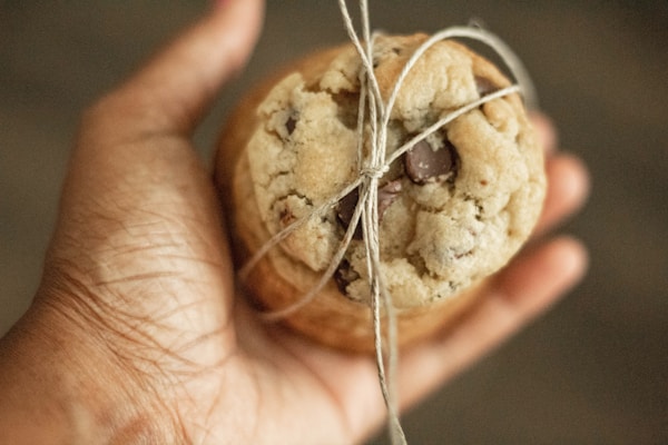 A hand is holding a chocolate chip cookie tied with a simple piece of string. The cookie appears to be homemade, with a soft texture and visible chocolate chunks.