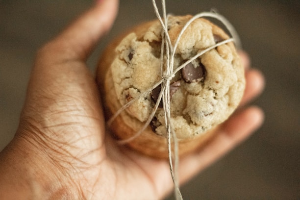 A hand is holding a chocolate chip cookie tied with a simple piece of string. The cookie appears to be homemade, with a soft texture and visible chocolate chunks.