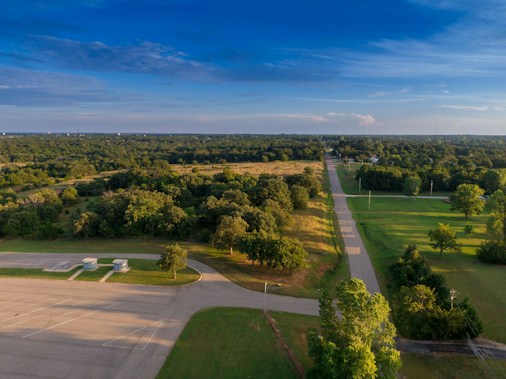 A rural landscape featuring a road running parallel to a dense cluster of trees and vegetation. The sky is clear with patches of white clouds, and the horizon is visible in the distance. There is an empty parking area with few structures, possibly utility installations, set amidst well-maintained grass.