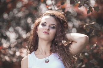 A woman with long, wavy hair poses outdoors against a background of blurred foliage. She has a confident expression, wearing a white top and a necklace. The lighting creates a soft, dreamy effect with bokeh circles in the background.