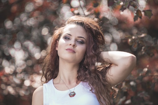 A woman with long, wavy hair poses outdoors against a background of blurred foliage. She has a confident expression, wearing a white top and a necklace. The lighting creates a soft, dreamy effect with bokeh circles in the background.