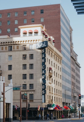 A multi-story building with a retro-style facade houses Hotel San Carlos. The architecture features a large vertical sign with ‘Hotel’ at the top and ‘San Carlos’ running downward. Several flags, including Mexican and Italian, are displayed along the street level. The building is complemented by adjacent modern high-rise buildings.