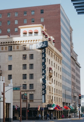 A multi-story building with a retro-style facade houses Hotel San Carlos. The architecture features a large vertical sign with &lsquo;Hotel&rsquo; at the top and &lsquo;San Carlos&rsquo; running downward. Several flags, including Mexican and Italian, are displayed along the street level. The building is complemented by adjacent modern high-rise buildings.