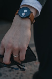 A person's hand wearing a wristwatch with a brown leather strap and a dark face featuring two subdials. The hand is holding a pair of black sunglasses and a small black wallet. Clothing includes a white shirt sleeve under a dark coat or sweater.