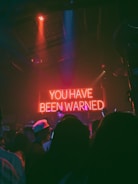 A dimly lit room with a crowd of people, mostly silhouetted, facing a prominent neon sign that reads 'YOU HAVE BEEN WARNED.' The atmosphere is that of an indoor venue or club with colorful lighting and shadows casting an enigmatic ambience.