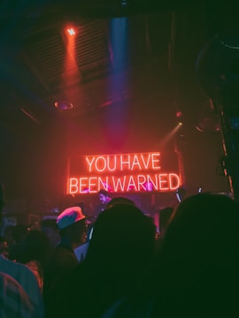 A dimly lit room with a crowd of people, mostly silhouetted, facing a prominent neon sign that reads 'YOU HAVE BEEN WARNED.' The atmosphere is that of an indoor venue or club with colorful lighting and shadows casting an enigmatic ambience.